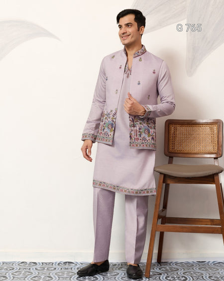 Man wearing a light purple traditional outfit with a jacket and kurta, standing against a white wall.