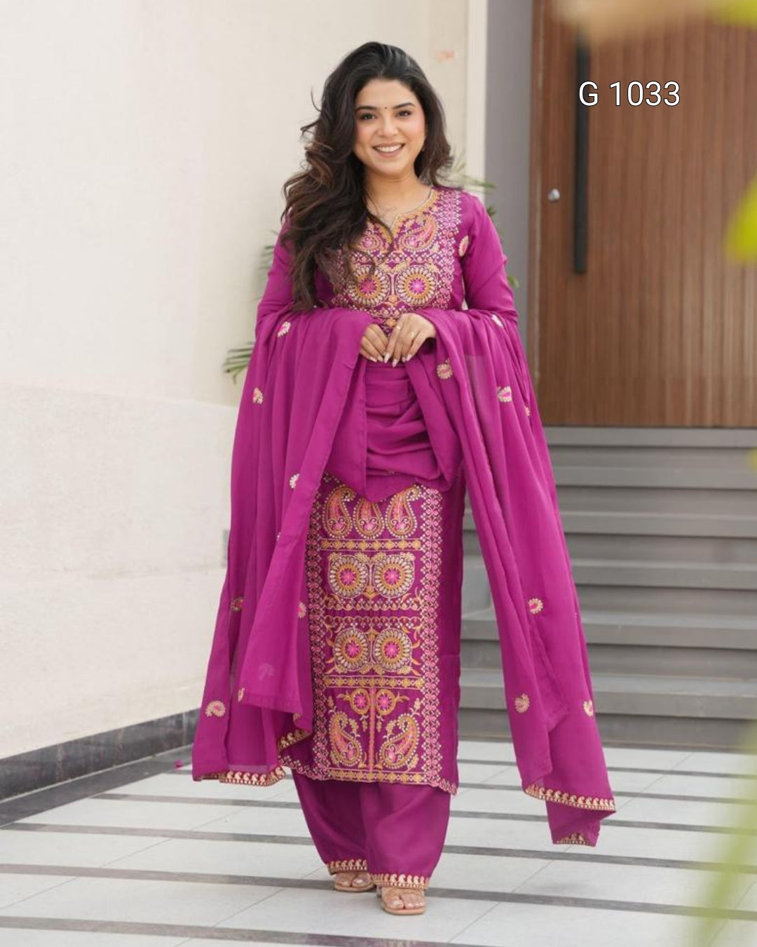 A woman modeling a magenta purple ethnic salwar kameez suit with intricate gold and multicolor embroidery, paired with matching embroidered trousers and a draped dupatta, set against an outdoor architectural background.