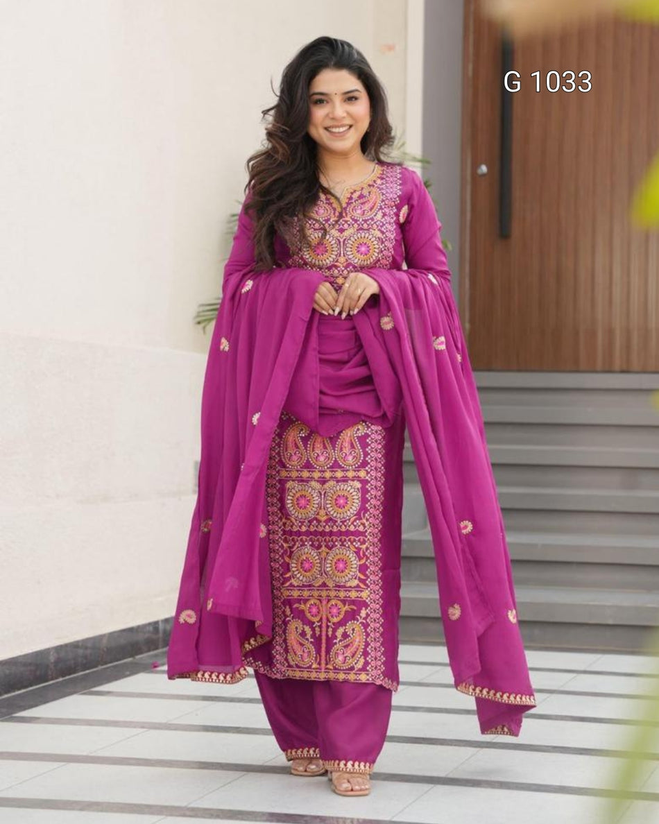 A woman modeling a magenta purple ethnic salwar kameez suit with intricate gold and multicolor embroidery, paired with matching embroidered trousers and a draped dupatta, set against an outdoor architectural background.