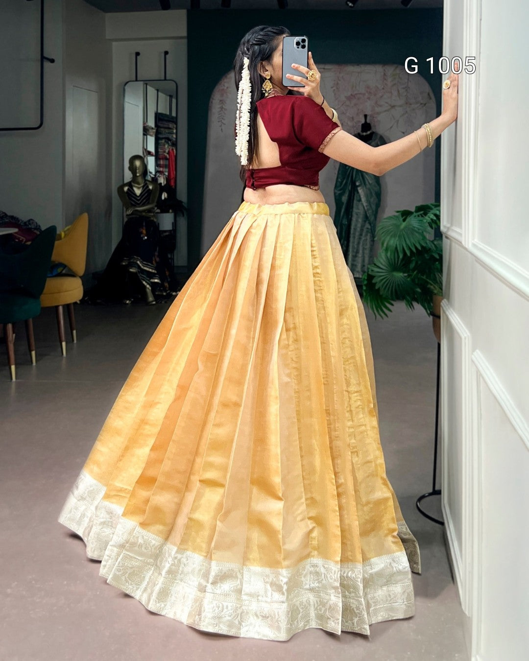 A woman wearing a traditional Indian outfit with a maroon short-sleeve crop top and a voluminous golden-yellow flared skirt with a white border, taking a mirror selfie in a boutique.