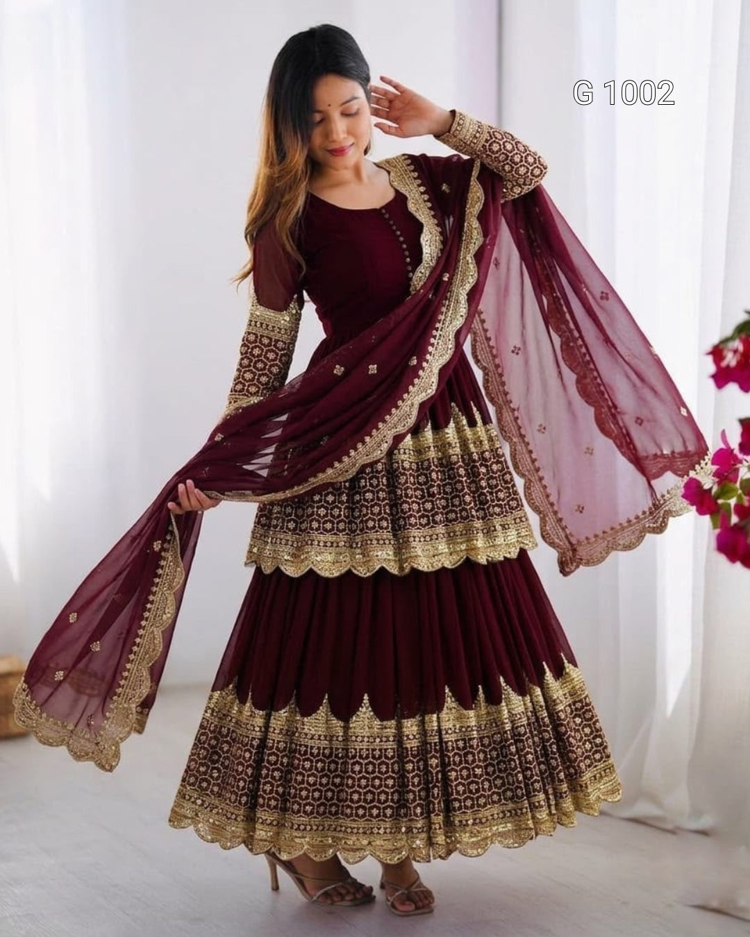 A woman models a deep maroon Anarkali gown with intricate gold embroidery and a matching dupatta, set against a soft white curtain background.