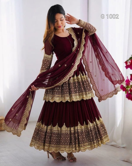A woman models a deep maroon Anarkali gown with intricate gold embroidery and a matching dupatta, set against a soft white curtain background.