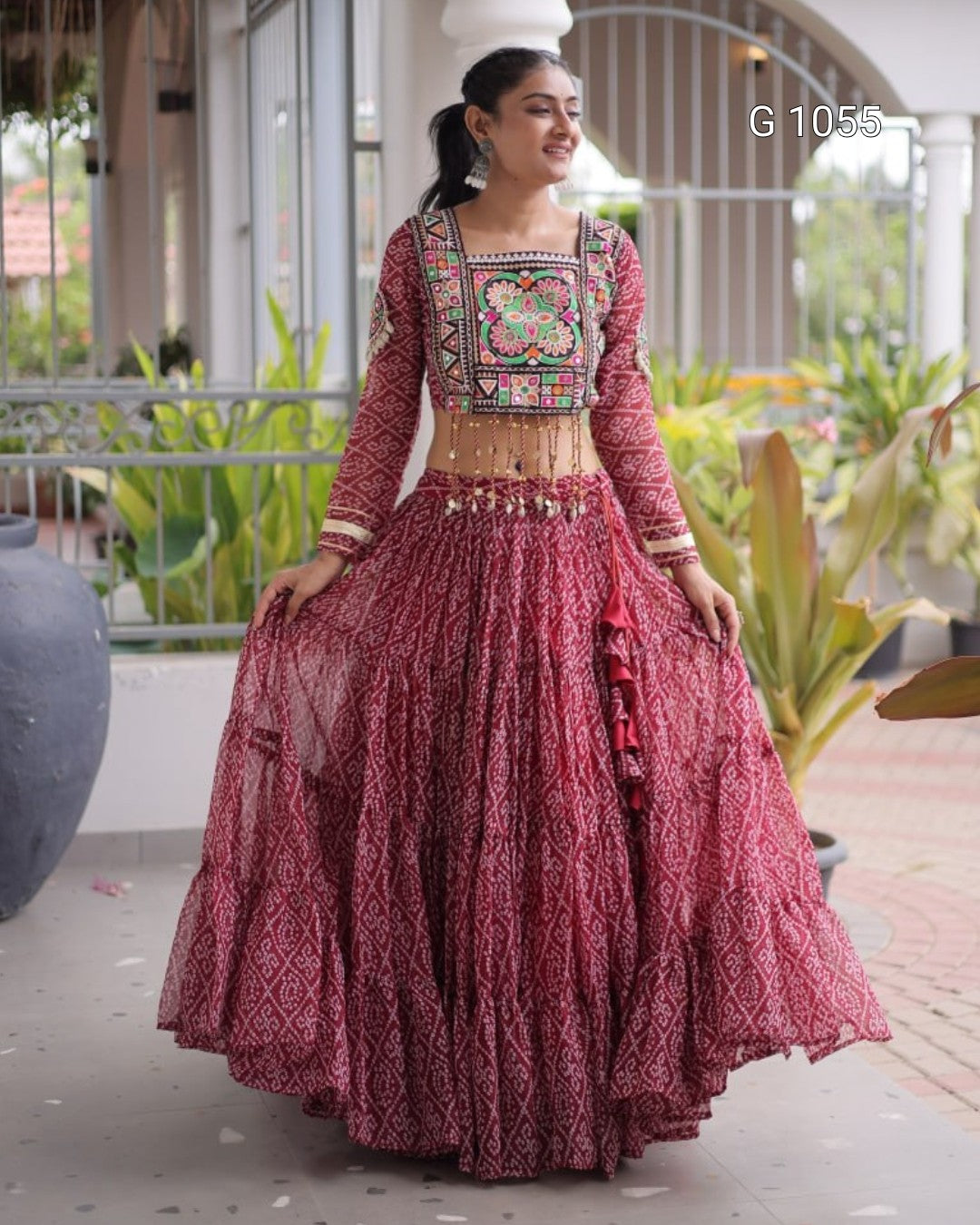 A woman wearing a traditional Indian lehenga choli outfit in deep maroon with intricate multicolor embroidery on the bodice and a heavily embellished flared skirt, standing in an outdoor patio setting.