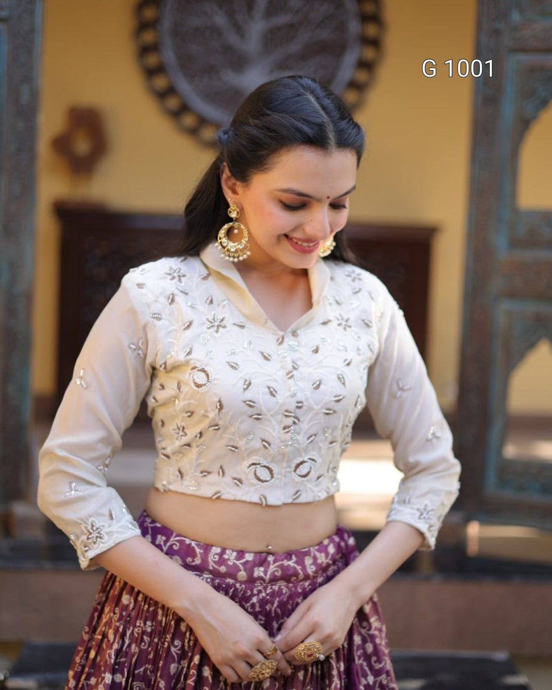 A woman wearing a cream-colored embroidered blouse with three-quarter sleeves and a purple patterned skirt, standing in an ornate indoor setting. The text 'G 1001' is visible in the top right corner.