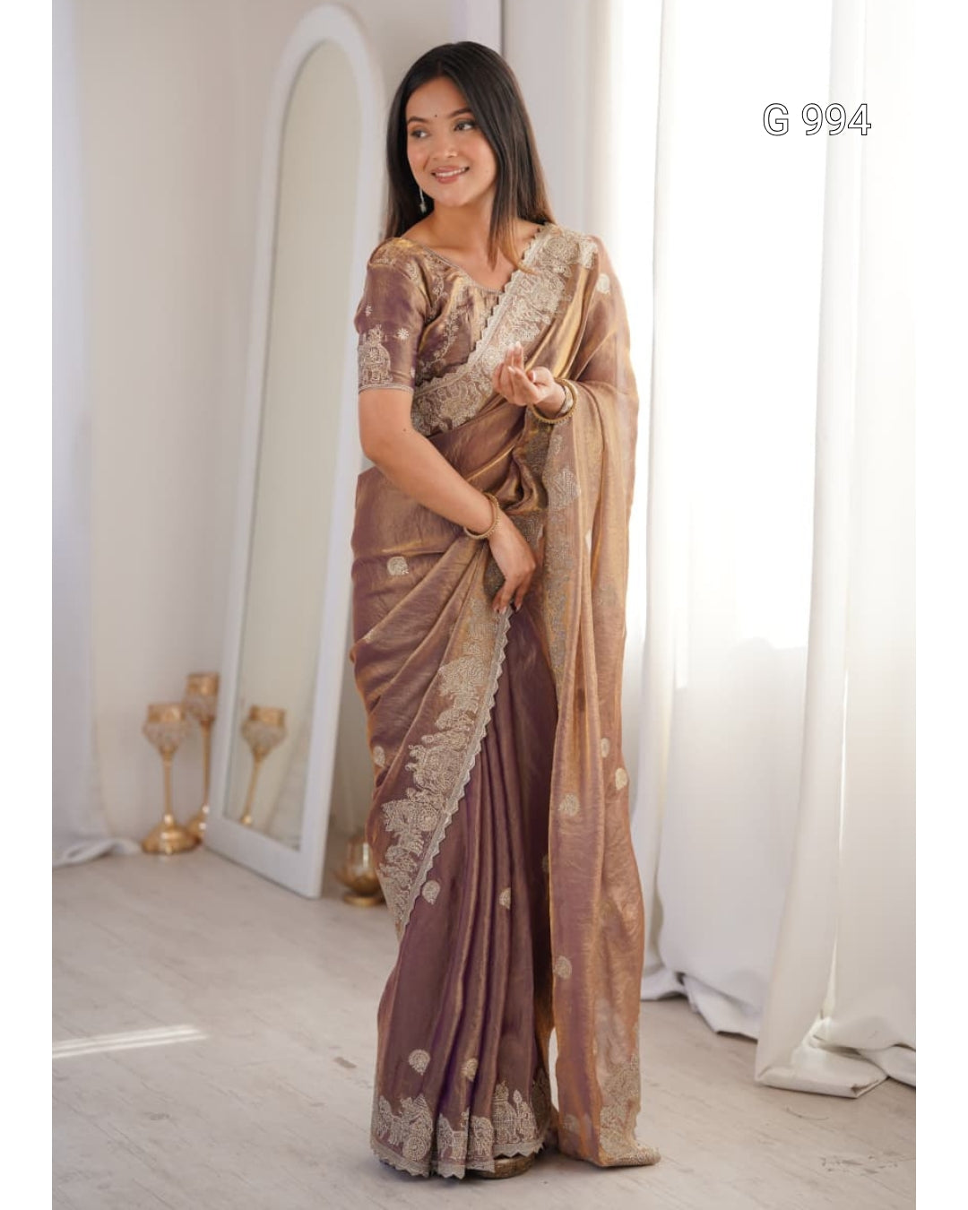 A woman models a traditional Indian saree in a bright indoor setting. The saree is a muted brownish-purple with intricate gold zari embroidery and motifs, paired with a matching embroidered blouse.