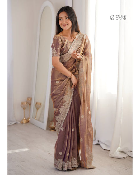 A woman models a traditional Indian saree in a bright indoor setting. The saree is a muted brownish-purple with intricate gold zari embroidery and motifs, paired with a matching embroidered blouse.