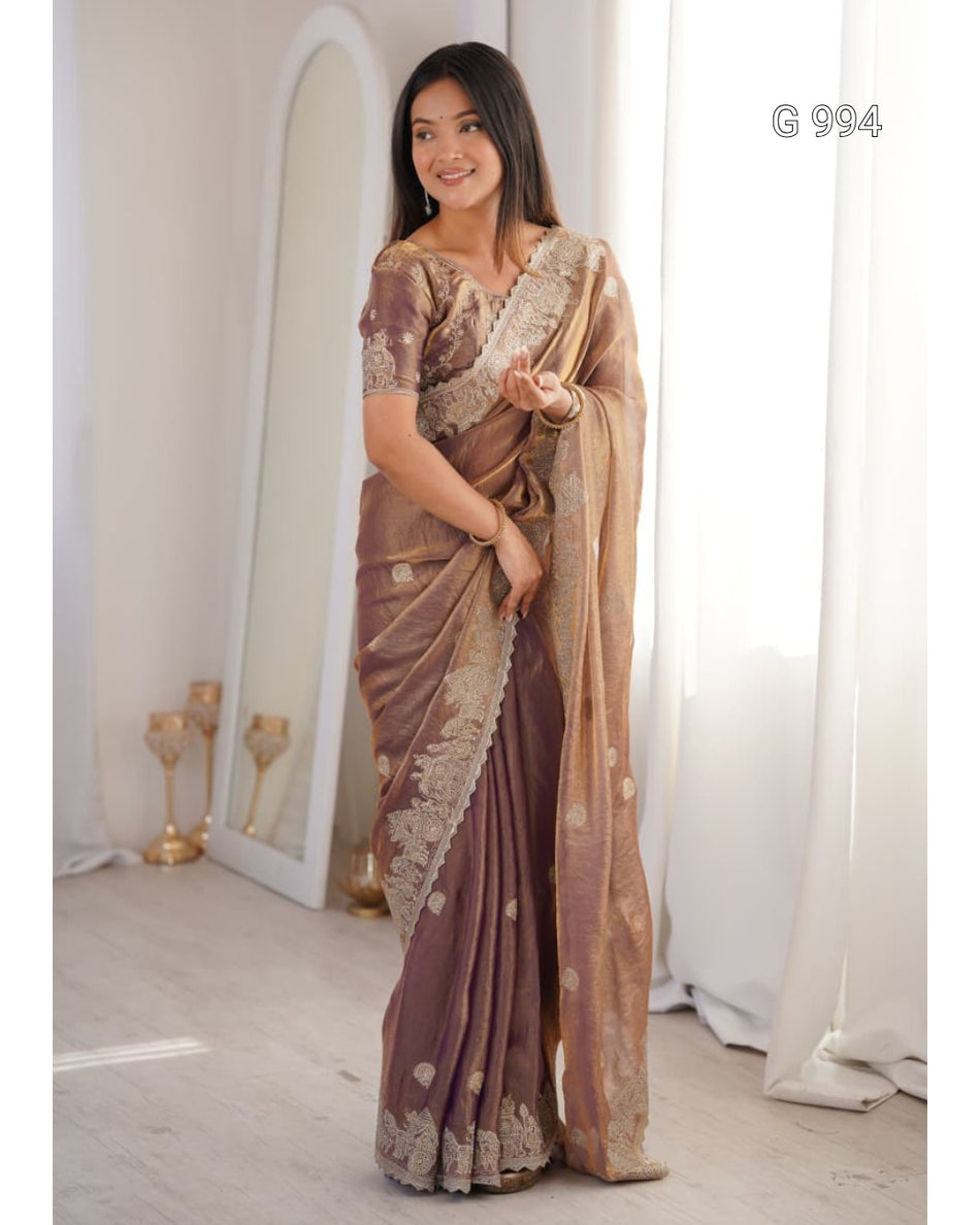 A woman models a traditional Indian saree in a bright indoor setting. The saree is a muted brownish-purple with intricate gold zari embroidery and motifs, paired with a matching embroidered blouse.
