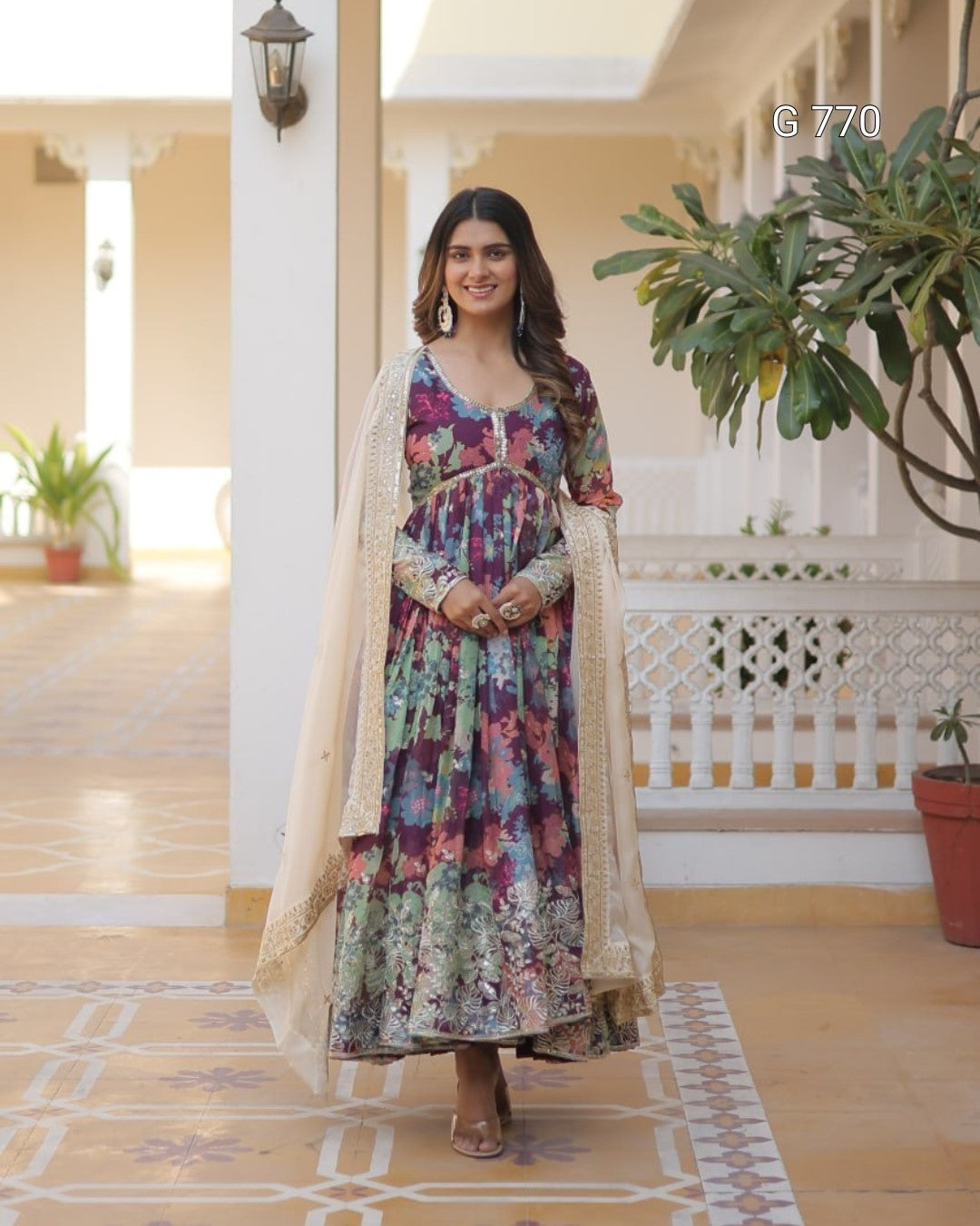 Woman in a floral dress standing in an outdoor setting with plants and architectural elements.