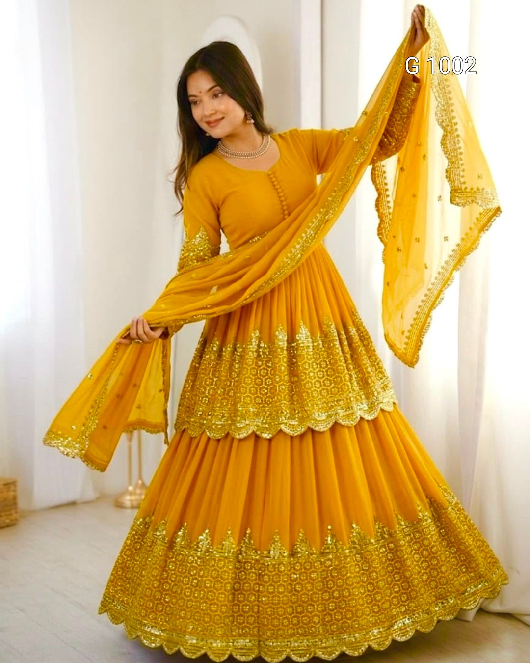 A woman modeling a traditional Indian outfit consisting of a bright yellow anarkali-style dress with gold embroidery, a matching long-sleeve blouse, and a sheer yellow dupatta with gold border detailing, set against a white curtain background.