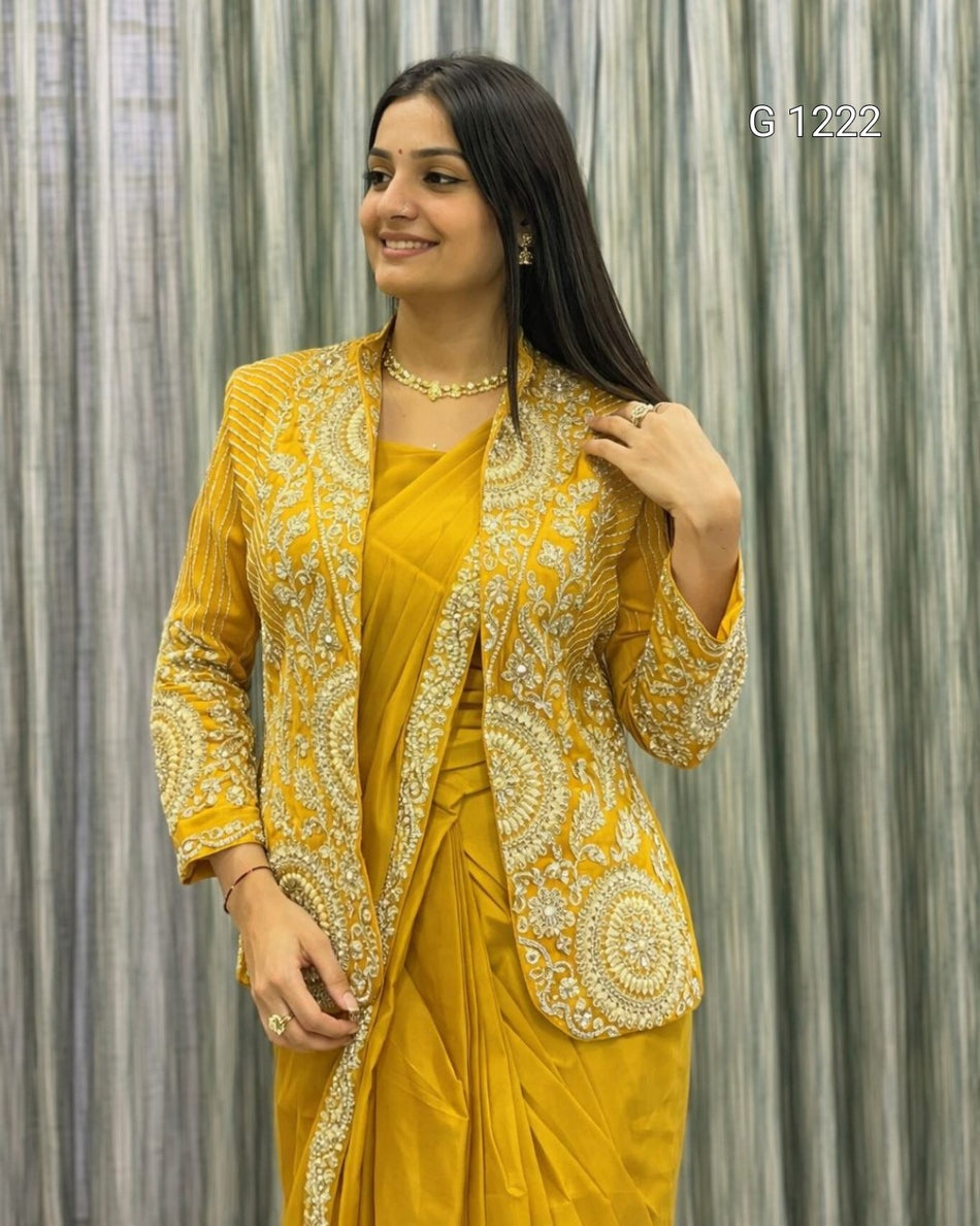 A woman modeling a mustard yellow traditional outfit consisting of a heavily embellished embroidered jacket and a matching draped skirt, posed against a gray textured curtain backdrop.