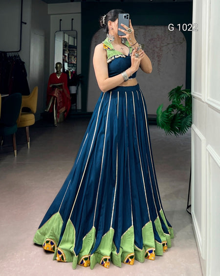 A woman takes a mirror selfie in a boutique wearing a traditional Indian outfit consisting of a dark teal blue skirt with vertical gold stripes and light green borders, paired with a matching sleeveless top. The background shows a clothing store interior with a mannequin and other garments.