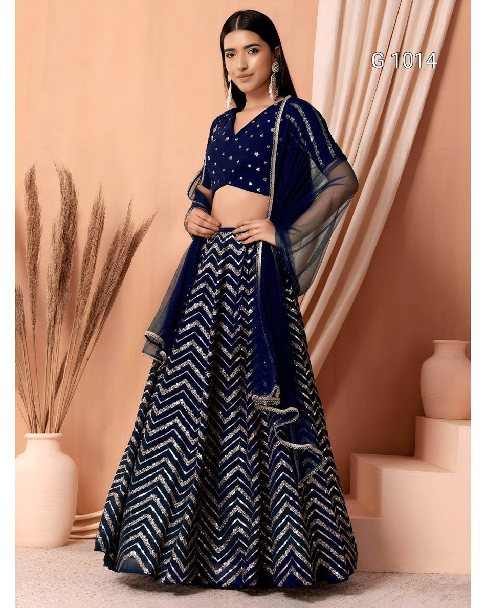 A woman models a navy blue and silver lehenga choli set with a matching dupatta in a peach-colored studio setting with decorative vases.