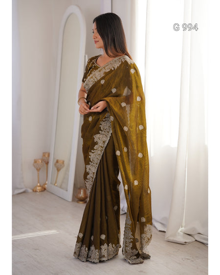 A woman modeling a dark olive green saree with beige floral embroidery and a matching embroidered blouse, standing in a bright room with a mirror and white curtains.