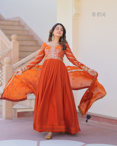 Woman in an orange traditional outfit with intricate designs, standing in a decorative indoor setting.