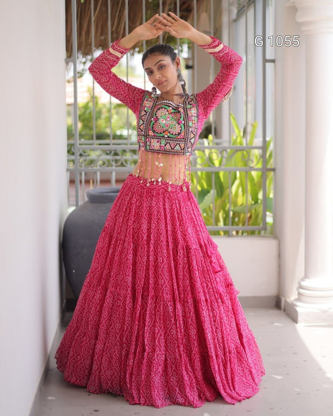 A woman modeling a bright pink traditional Indian lehenga choli set with intricate embroidery on the bodice, standing in a covered outdoor walkway.