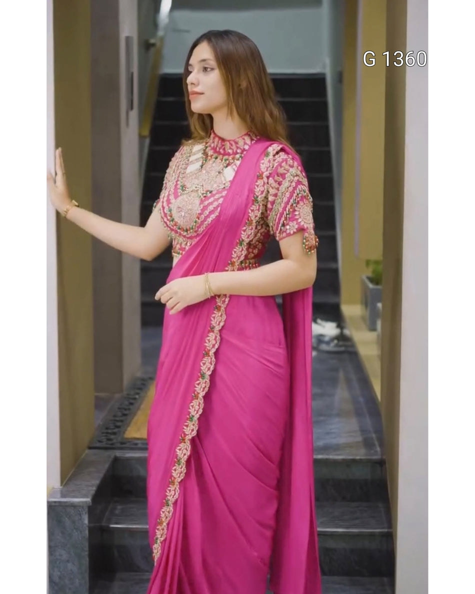 A woman wearing a bright pink saree with gold and silver embroidery on the blouse and border, standing in an indoor hallway with dark stairs in the background.