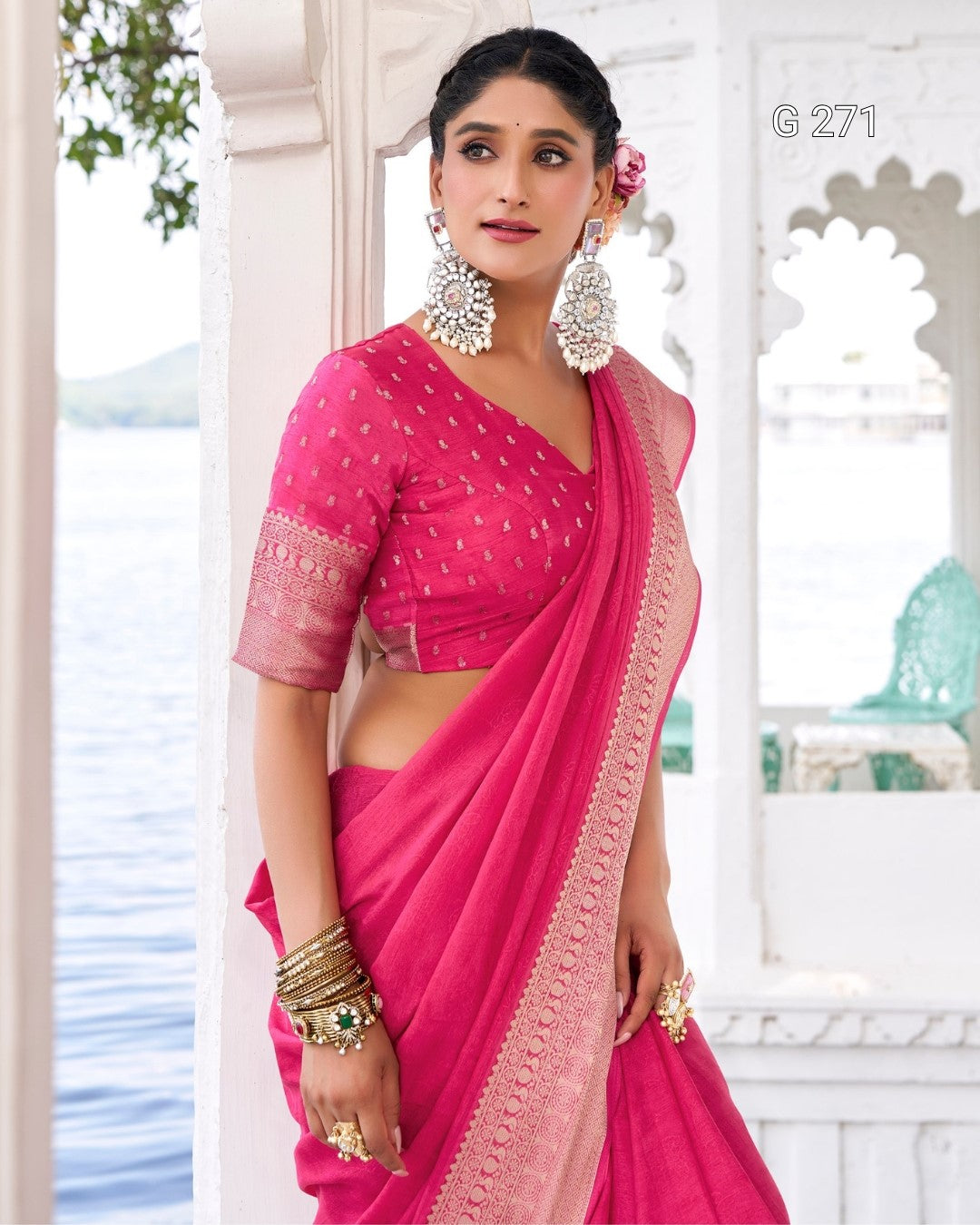 A woman modeling a bright pink saree with a matching embroidered blouse, standing in front of a white architectural background with water visible outside.