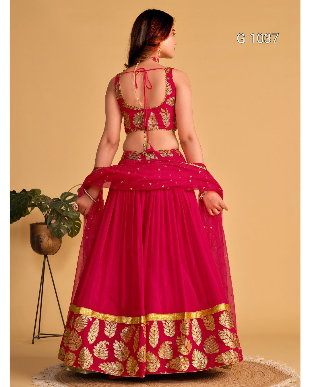 A woman modeling a bright pink lehenga choli set from the back, featuring gold leaf embroidery, a solid pink skirt, and a matching sheer dupatta, set against a warm beige background.