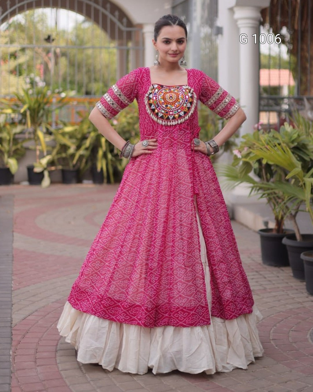 A woman modeling a bright pink Anarkali gown with intricate embroidery and a cream-colored ruffled underskirt, standing in an outdoor courtyard setting.