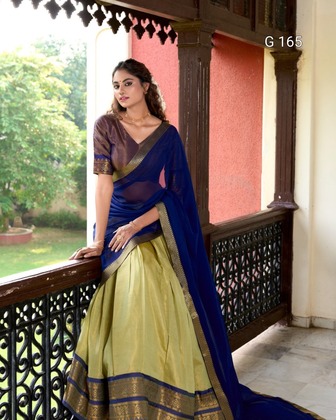 Woman in a blue and green saree standing on a balcony with a garden view.