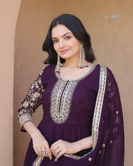 A woman modeling a deep purple traditional Indian salwar kameez set with intricate silver embroidery and a matching sheer dupatta, posed against a warm beige stucco wall.