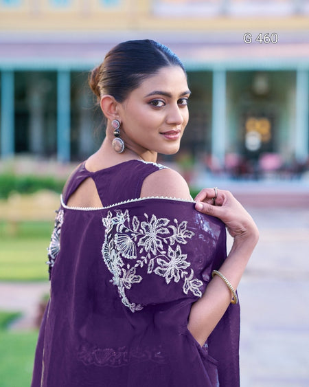 A woman wearing a deep purple off-the-shoulder kurta with white floral embroidery, photographed outdoors with a blurred building background.