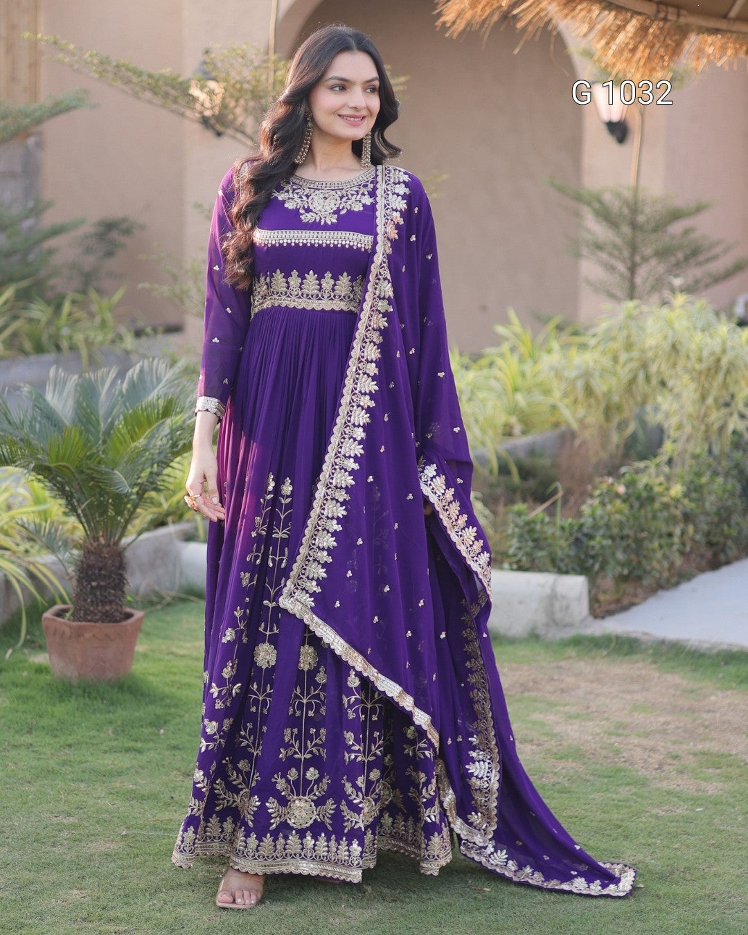 A woman stands outdoors on a lawn wearing a purple traditional outfit with extensive gold embroidery and a matching draped scarf. The setting includes grass, plants, and a building in the background.