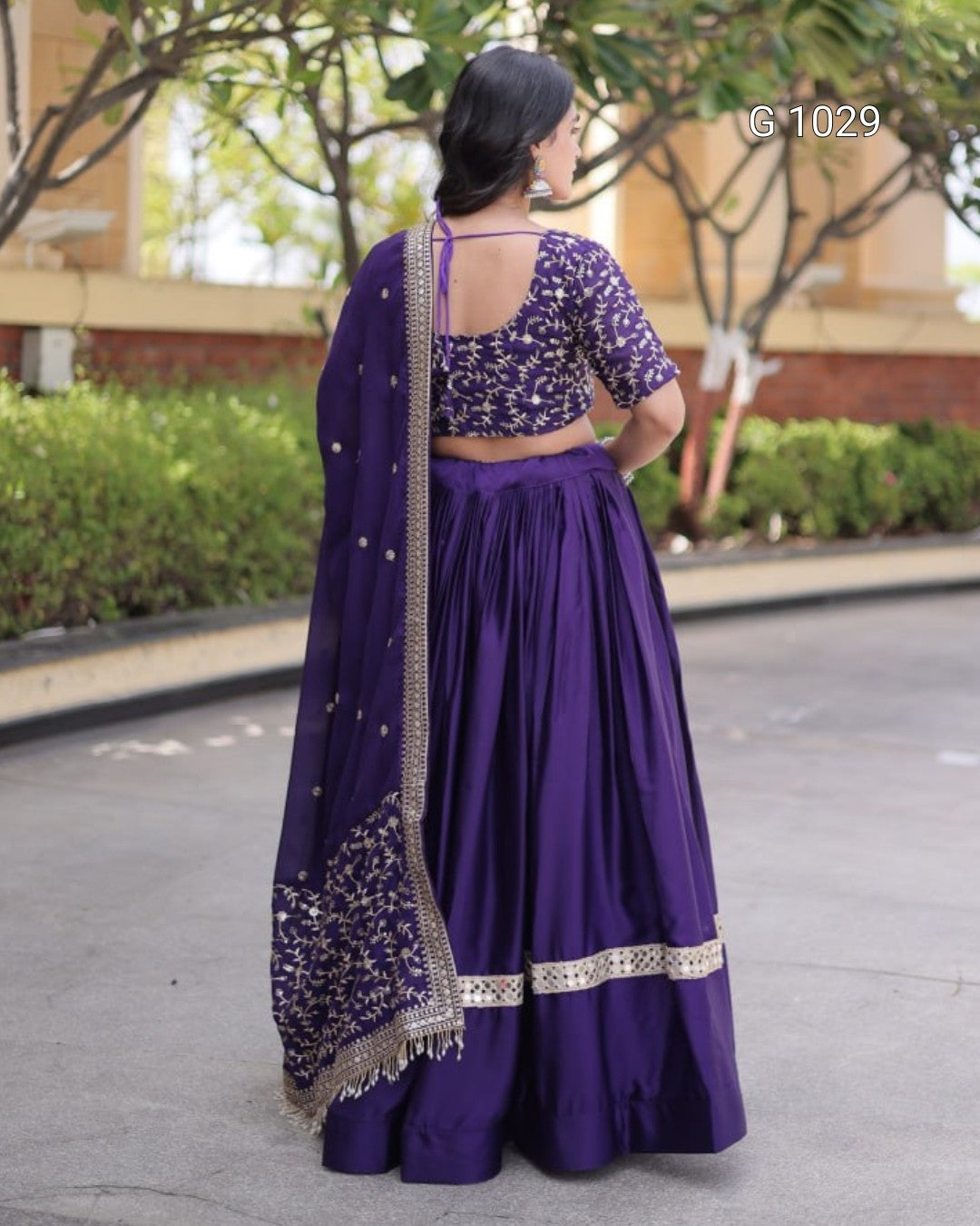 A woman stands outdoors facing away from the camera, wearing a deep purple traditional outfit with a heavily embroidered purple blouse, matching pleated skirt, and a sheer embroidered dupatta.