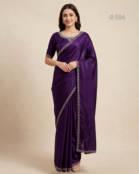 A woman modeling a deep purple silk saree with silver embroidered borders and a matching purple blouse, set against a plain beige studio background.