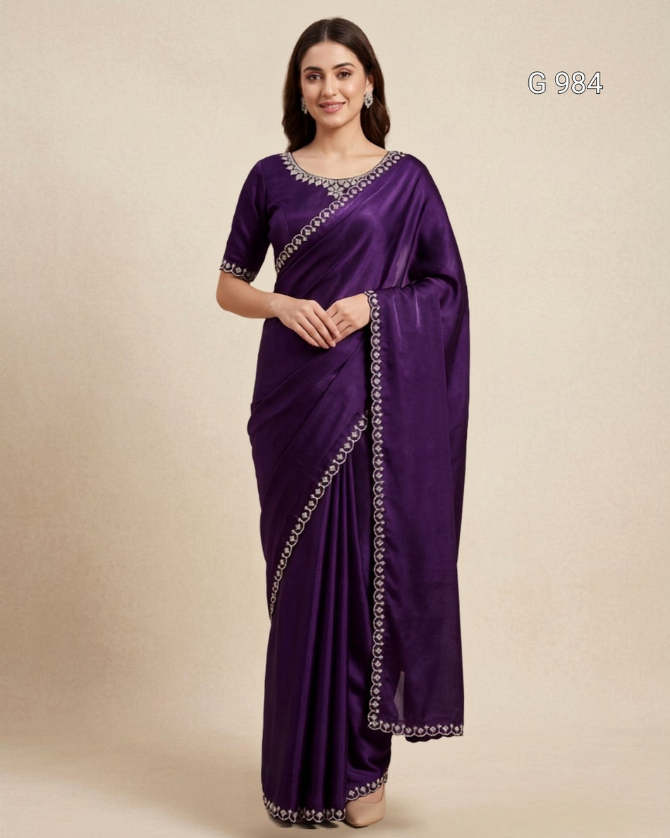 A woman modeling a deep purple silk saree with silver embroidered borders and a matching purple blouse, set against a plain beige studio background.