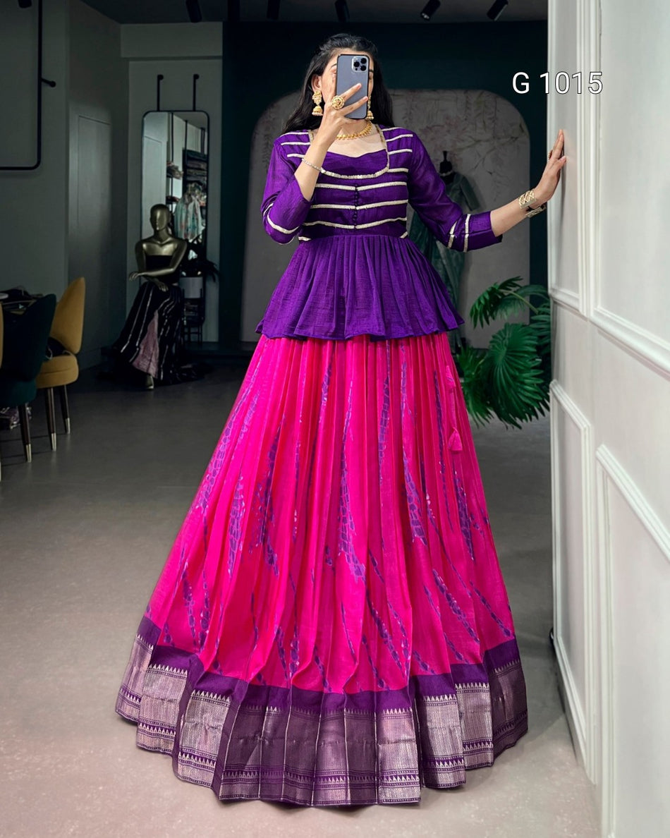 A woman takes a mirror selfie wearing a traditional Indian ethnic outfit consisting of a purple peplum top with gold stripes and a bright pink flared skirt with purple borders and subtle metallic patterns.