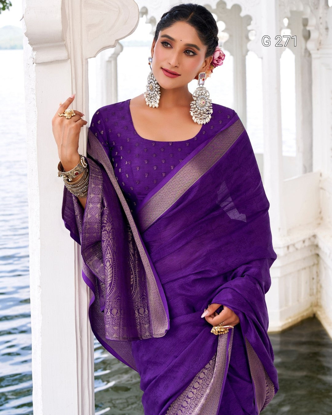 A woman models a purple saree with a matching embroidered blouse, draped over a white architectural porch with water visible in the background.