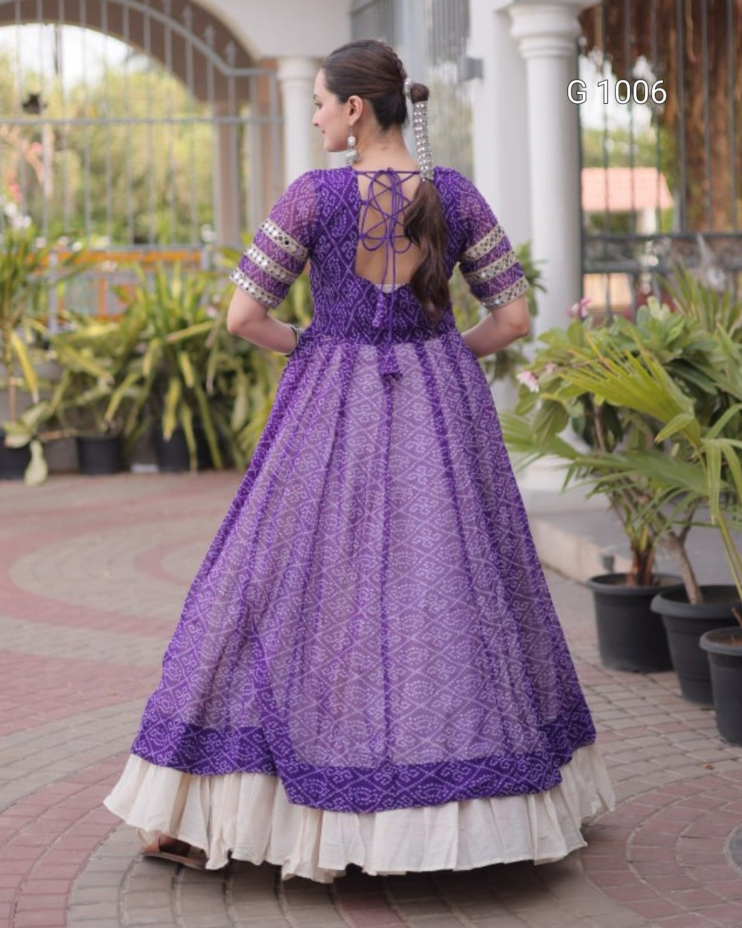 A woman wearing a purple embroidered Anarkali gown, shown from the back in an outdoor courtyard setting with potted plants.