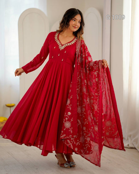 A woman modeling a long red anarkali-style dress with silver embroidery and a matching sheer red dupatta draped over one arm, standing in a bright indoor setting with white arches and curtains.