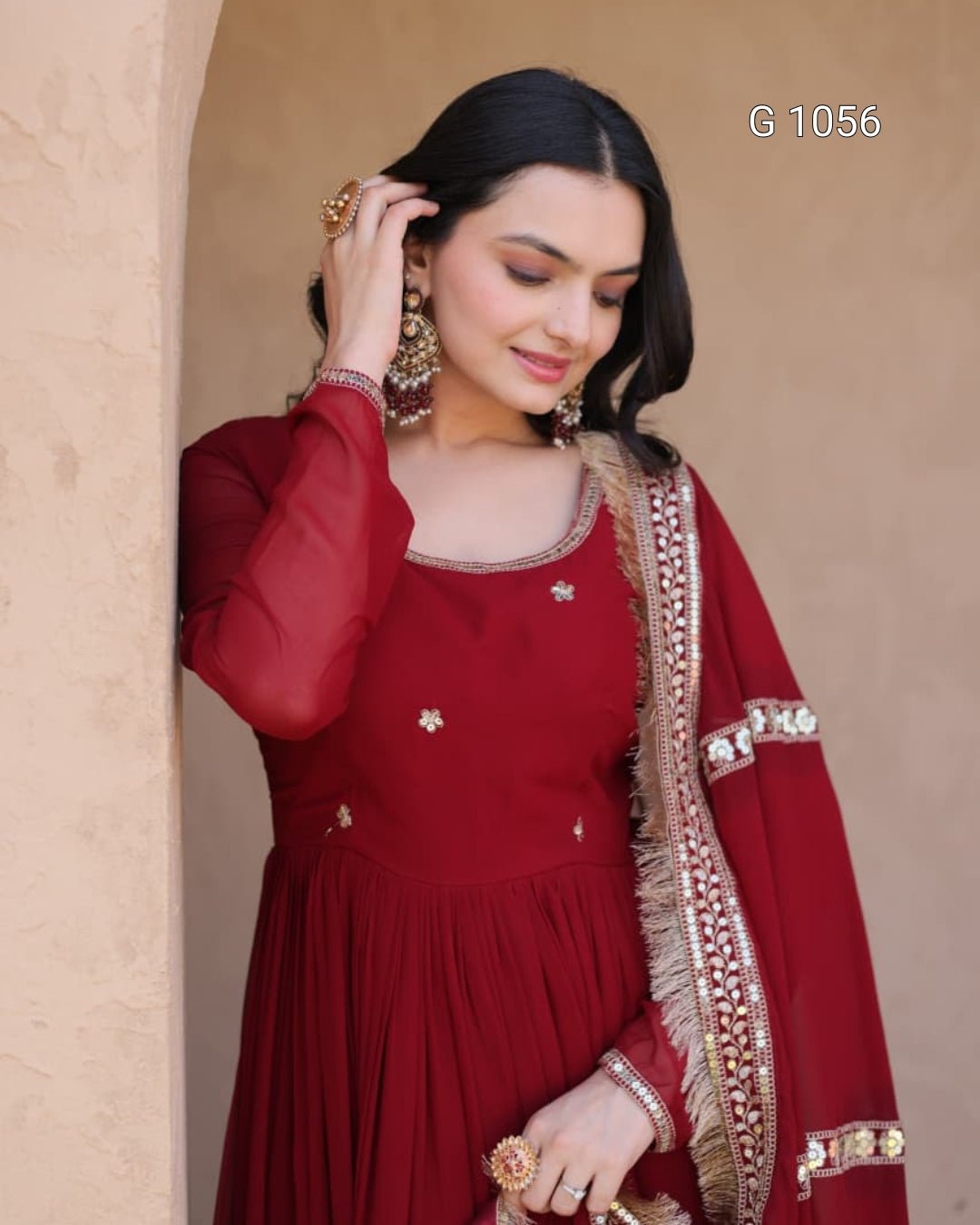 A woman wearing a deep red Anarkali gown with gold embroidery and a matching dupatta draped over her shoulder, posed against a beige stucco wall.