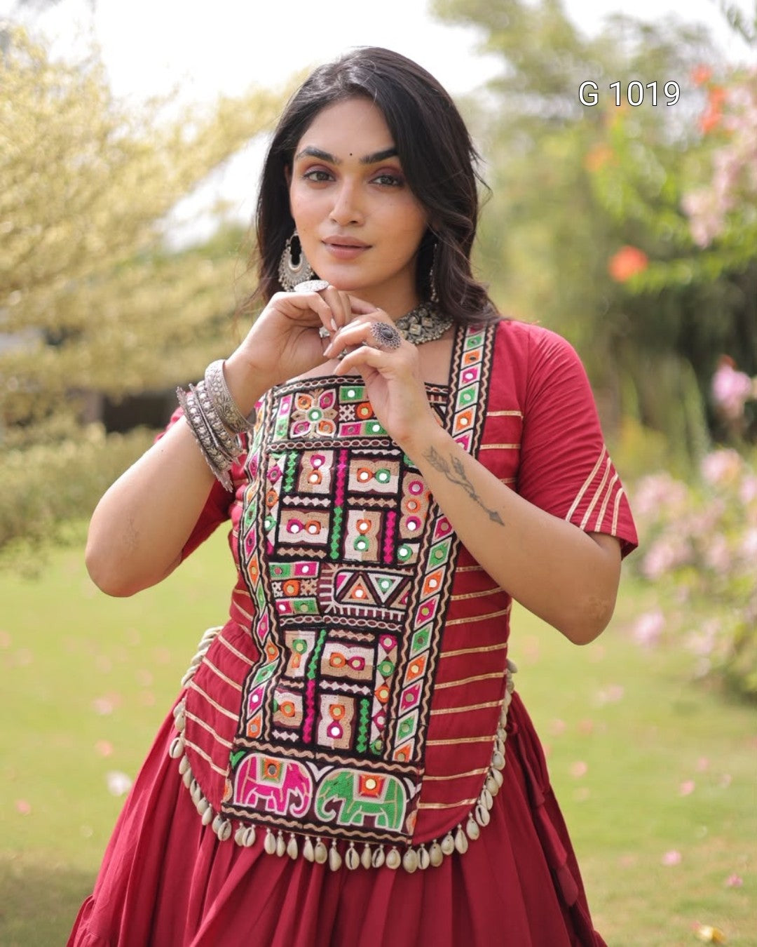 A woman wearing a red traditional embroidered dress with colorful geometric mirror work on the bodice and elephant motifs near the hem, standing outdoors in a garden setting.