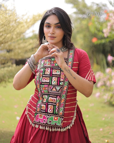 A woman wearing a red traditional embroidered dress with colorful geometric mirror work on the bodice and elephant motifs near the hem, standing outdoors in a garden setting.
