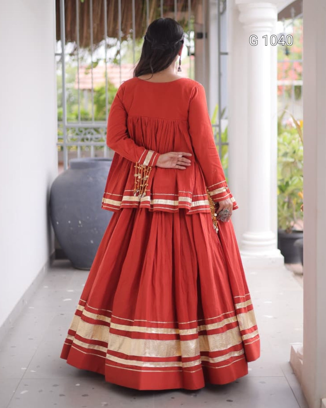 A woman viewed from behind wearing a red traditional long dress with beige and white striped borders, standing in a covered walkway.