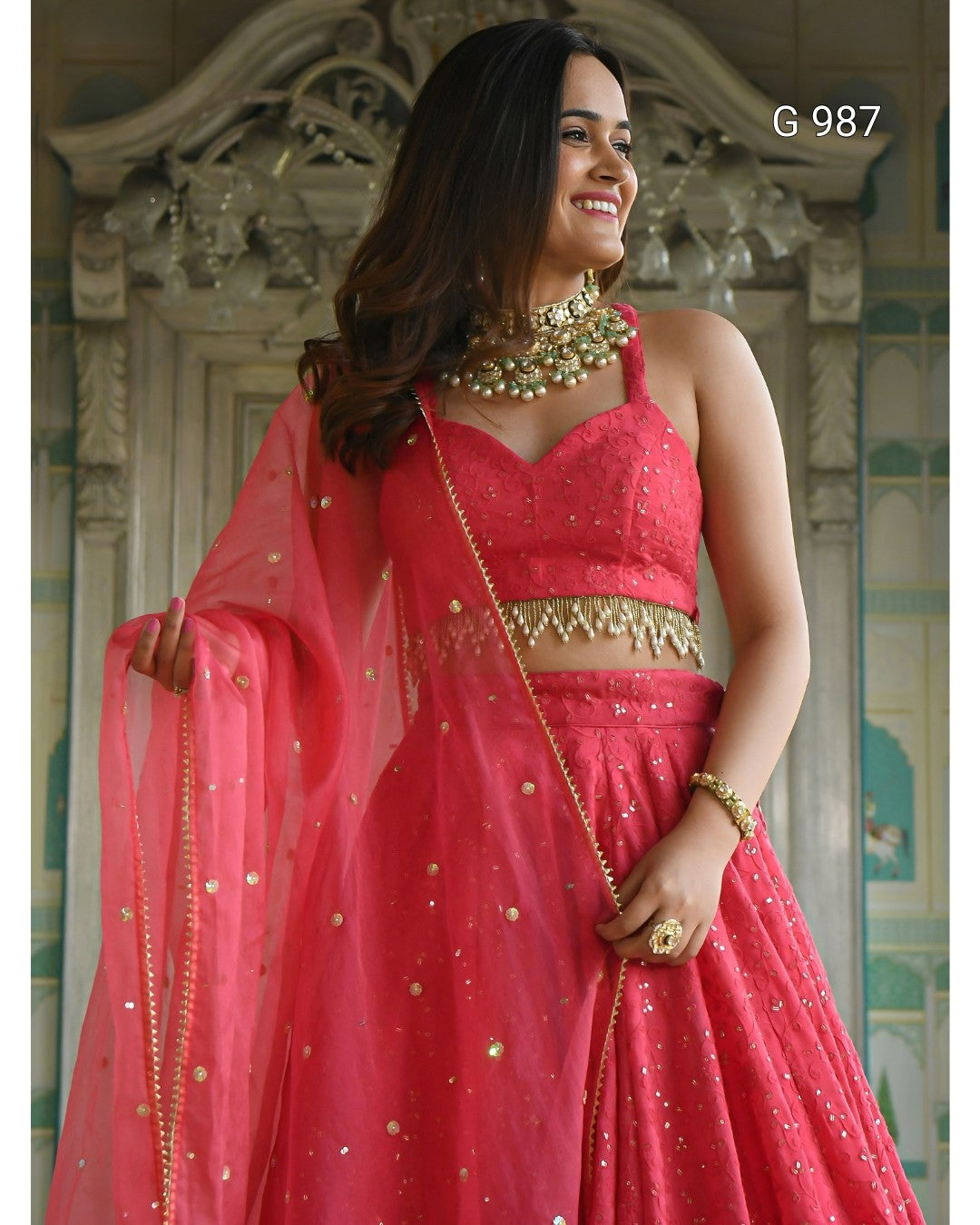A woman modeling a bright pink lehenga choli set with gold embellishments, including a matching sheer dupatta, against an ornate classical background.