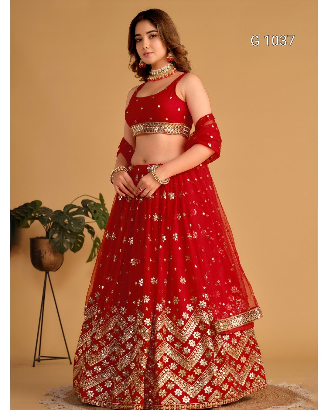 A woman models a red and gold embroidered Indian ethnic wear set, featuring a red off-the-shoulder lehenga skirt with gold floral embroidery and a matching red crop top. The set is photographed against a solid beige background with a small potted plant on the left.