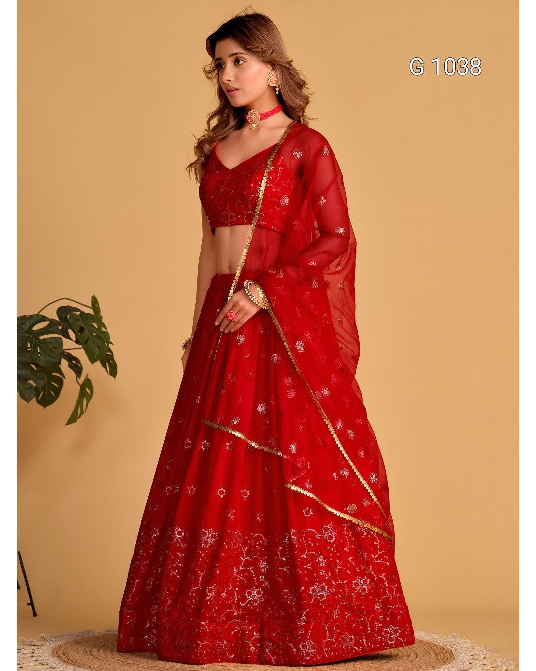 A woman models a red Indian ethnic lehenga choli suit with floral embroidery and gold borders, set against a solid beige background.