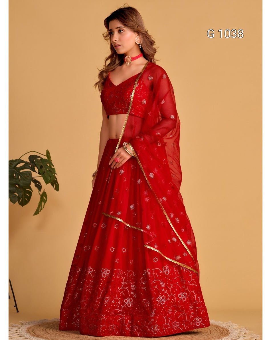 A woman models a red Indian ethnic lehenga choli suit with floral embroidery and gold borders, set against a solid beige background.