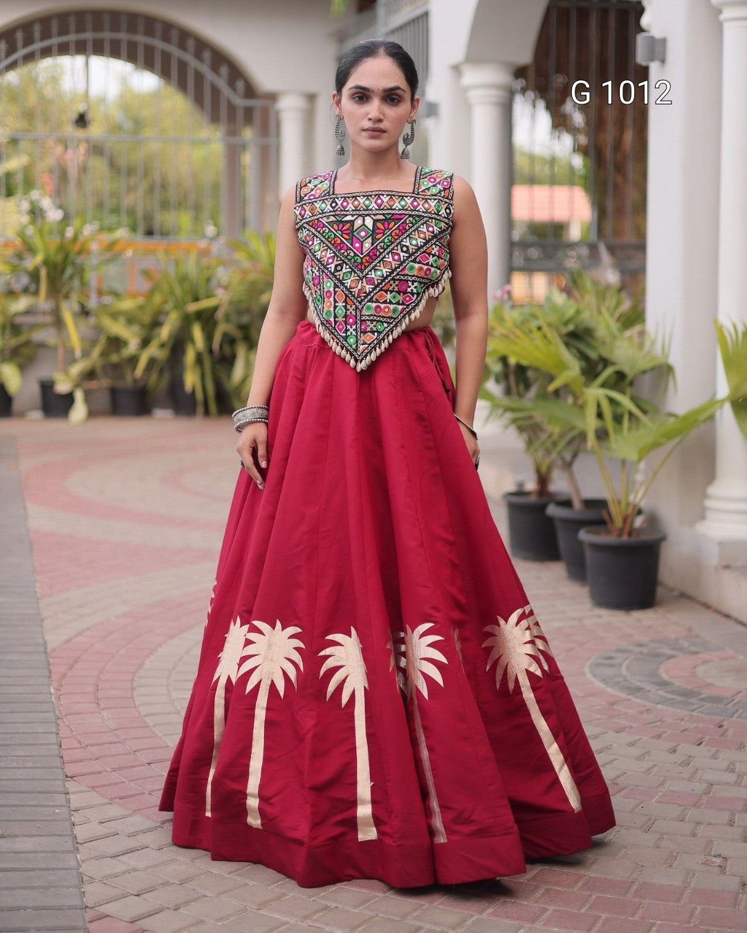 A woman models a traditional Indian anarkali gown with a deep red skirt featuring cream palm tree prints and a heavily embroidered multicolor bodice, set against an outdoor architectural background with potted plants.