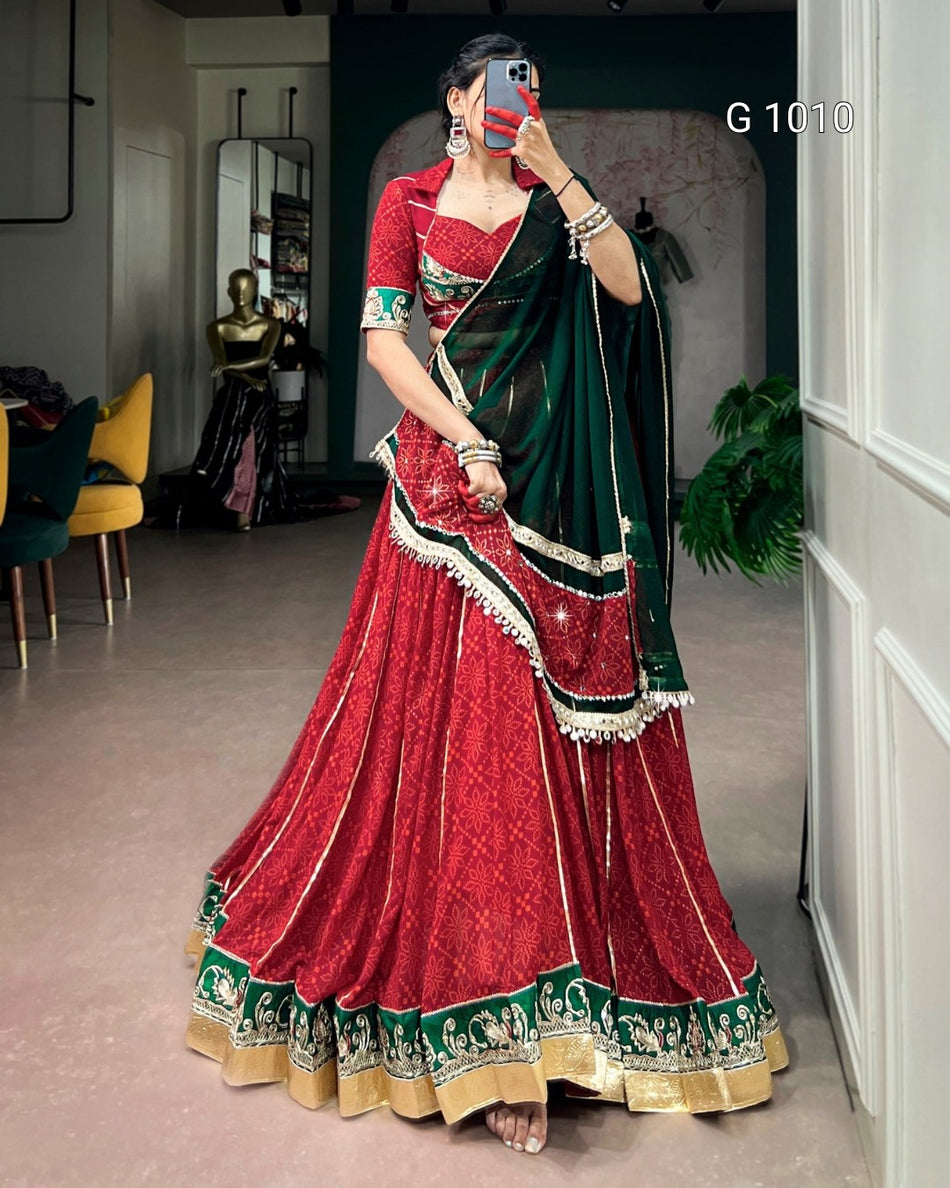 A woman modeling a traditional Indian lehenga choli set in a mirror selfie. The outfit features a red skirt with intricate gold and green border patterns, a red embroidered blouse, and a dark green dupatta draped over her shoulder.