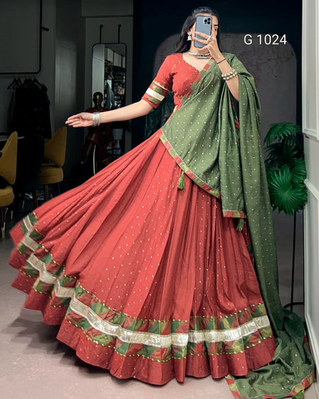 A woman modeling a traditional Indian ethnic outfit consisting of a rust-red flared skirt (lehenga) with a green and gold border, a matching rust-red blouse with green and gold trim, and a green dupatta with gold dot patterns. She is taking a mirror selfie in an indoor setting.