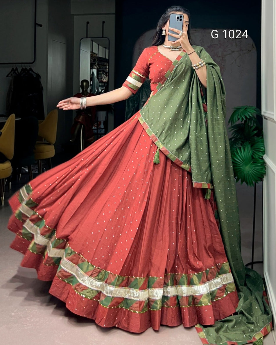 A woman modeling a traditional Indian ethnic outfit consisting of a rust-red flared skirt (lehenga) with a green and gold border, a matching rust-red blouse with green and gold trim, and a green dupatta with gold dot patterns. She is taking a mirror selfie in an indoor setting.