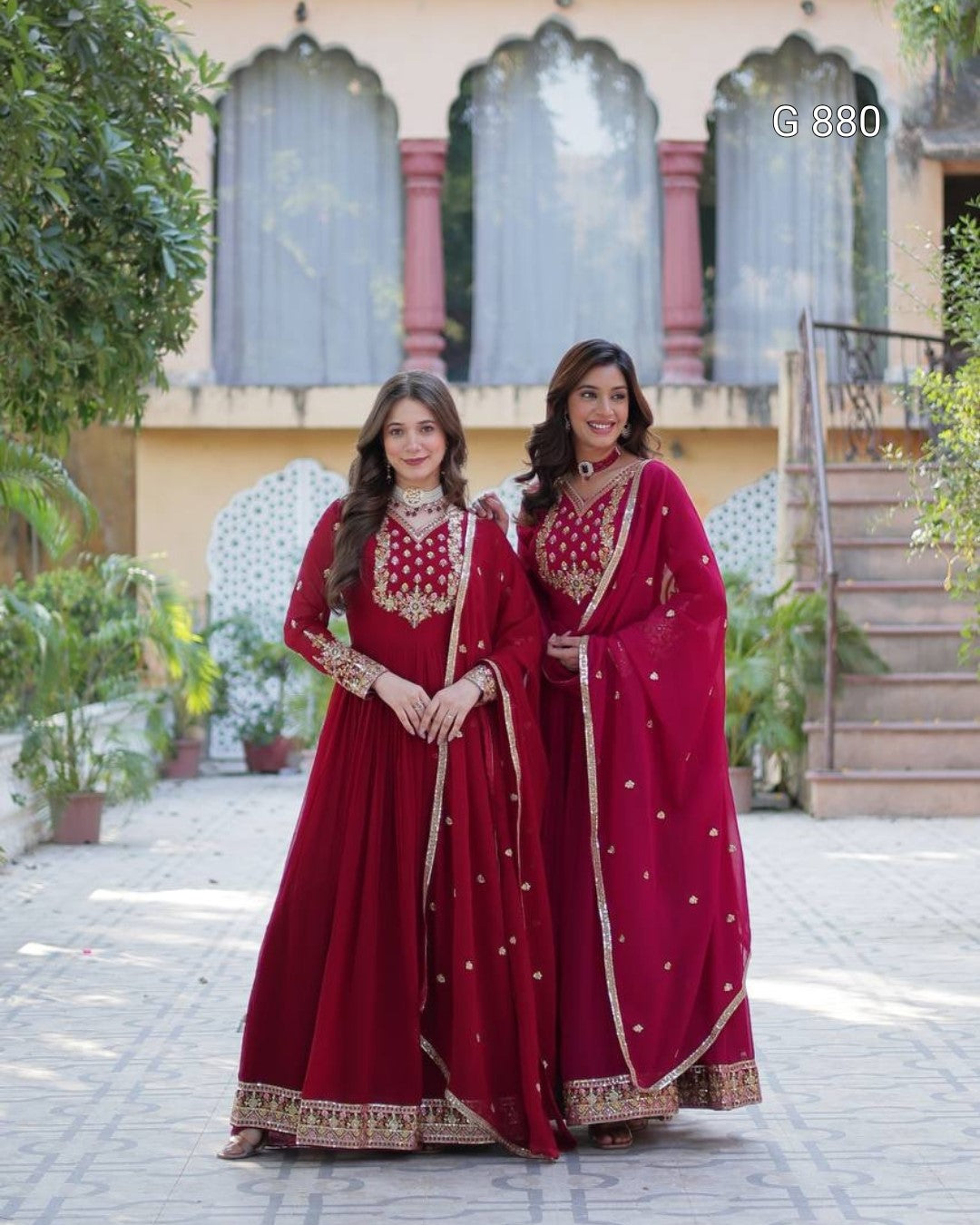 Two women in red traditional outfits standing outdoors with a building and plants in the background.