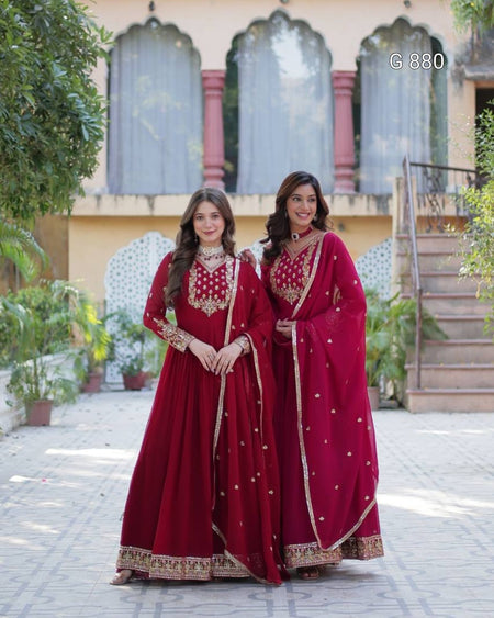 Two women in red traditional outfits standing outdoors with a building and plants in the background.