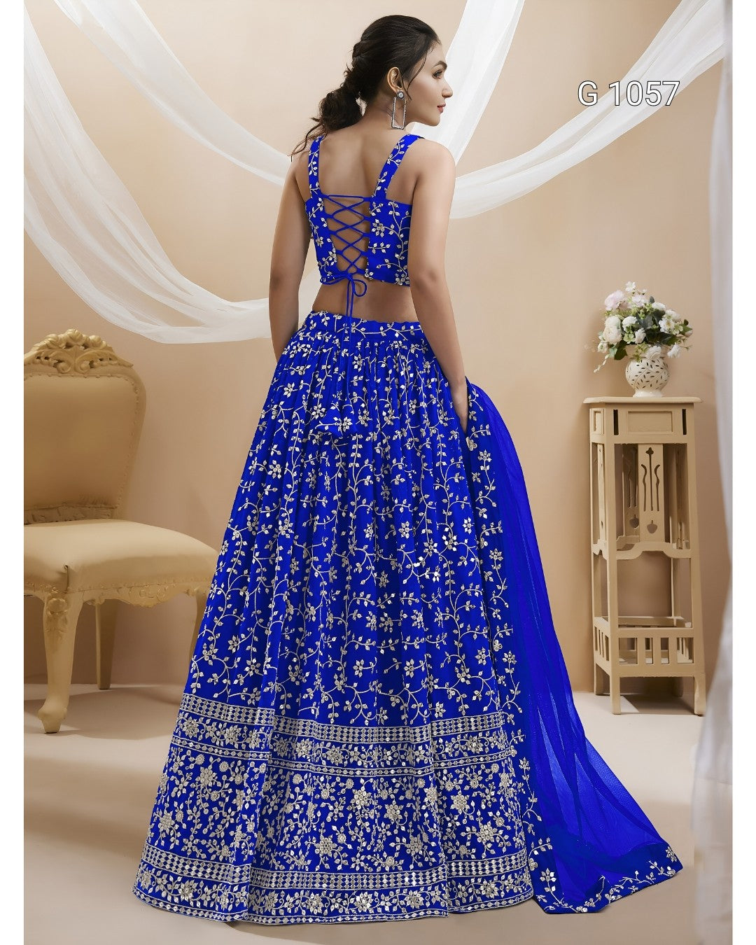 A woman wearing a royal blue lehenga choli with intricate silver and white floral embroidery, shown from the back in a decorated indoor setting.