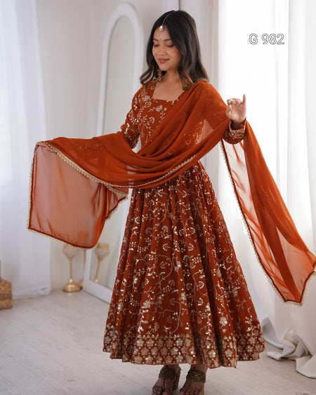 A woman models a rust-orange embroidered Anarkali gown with a matching sheer dupatta in a bright, white-walled room. The dress features intricate gold and silver floral embroidery, and the dupatta has gold border detailing.